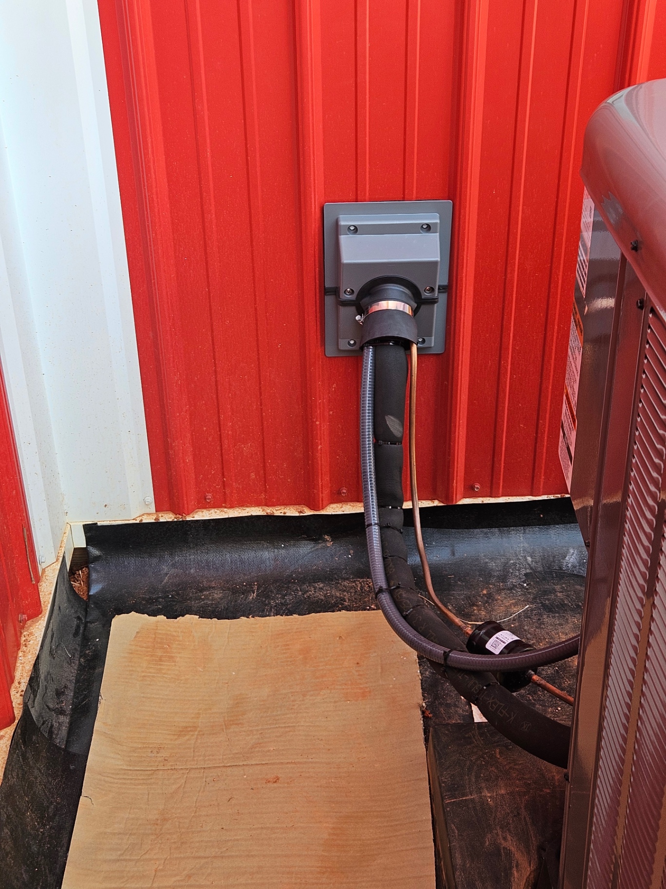 Insulated refrigerant lines and wiring entering a red metal building beside an outdoor HVAC unit